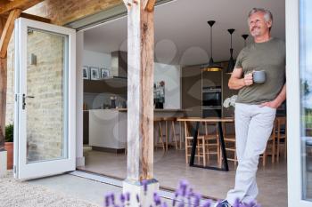 Senior Man Standing And Looking Out Of Kitchen Door Drinking Coffee