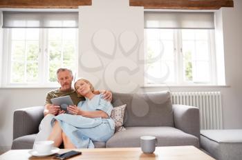 Senior Couple Sitting On Sofa In Lounge At Home Using Digital Tablet Together