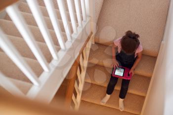 Overhead Shot Of Girl Sitting On Stairs At Home Playing With Digital Tablet
