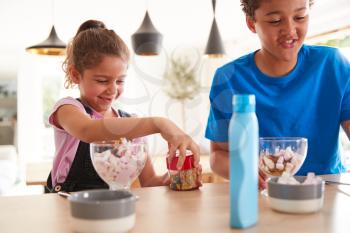 Children In Kitchen At Home Adding Sprinkles And Sauce To Ice Cream Dessert