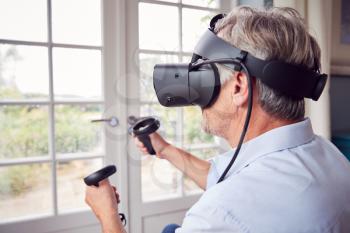 Mature Disabled Man In Wheelchair At Home Wearing Virtual Reality Headset Holding Gaming Controllers