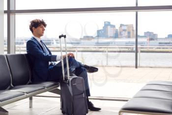 Businessman With Luggage Sitting In Airport Departure Lounge Looking Out Of Window