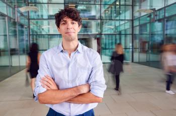 Portrait Of Young Businessman With Crossed Arms Standing In Lobby Of Busy Modern Office