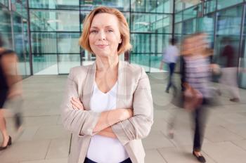 Portrait Of Mature Businesswoman With Crossed Arms Standing In Lobby Of Busy Modern Office