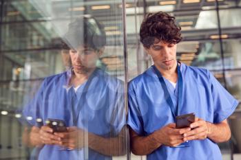 Young Male Doctor Wearing Scrubs Using Mobile Phone Leaning Against Wall In Modern Hospital Building