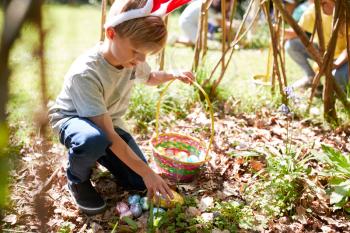 Group Of Children Wearing Bunny Ears Finding Easter Eggs Hidden In Garden