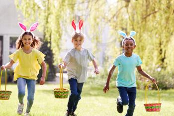 Group Of Children Wearing Bunny Ears Running On Easter Egg Hunt In Garden