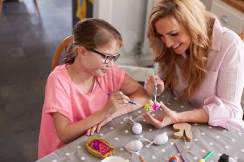 Mother With Daughter Sitting At Table Decorating Eggs For Easter At Home