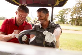 Two Mature Men Playing Golf Marking Scorecard In Buggy Driving Along Course