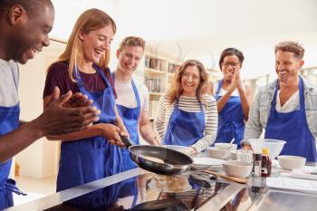 Female Teacher Making Pancake On Cooker In Cookery Class As Adult Students Look On