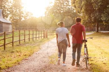 Rear View Shot Of Romantic Couple Walking And Pushing Bike Along Country Lane At Sunset