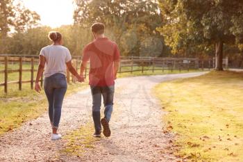 Rear View Of Romantic Couple Walking Hand In Hand Along Country Lane At Sunset