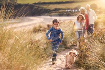 Grandchildren And Pet Dog Exploring Sand Dunes With Grandparents On Winter Beach Vacation