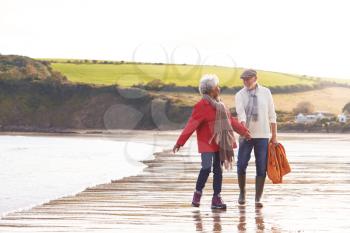 Senior Couple Hold Hands As They Walk Along Shoreline On Winter Beach Vacation