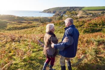 Rear View Of Loving Senior Couple Arm In Arm As They Walk Along Coast Path