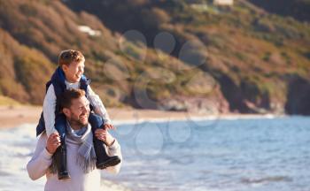 Father Giving Son Ride On Shoulders As They Walk Along Beach By Sea