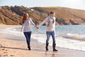 Loving Couple Holding Hands As They Walk Along Shoreline Of Beach Through Waves
