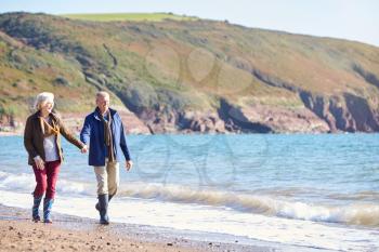 Loving Senior Couple Holding Hands As They Walk Along Shoreline Of Beach By Waves