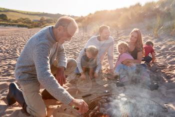 Grandfather Cooking As Multi-Generation Family Having Evening Barbecue Around Fire On Beach Vacation
