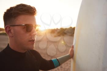 Young Man In Sunglasses Wearing Wetsuit Enjoying Surfing Staycation On Beach As Sun Sets