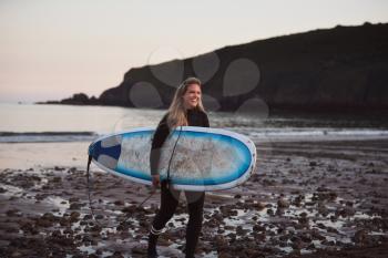 Woman Wearing Wetsuit Carrying Surfboard As She Walks Out Of Sea