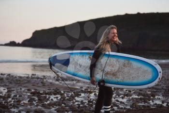 Woman Wearing Wetsuit Carrying Surfboard As She Walks Out Of Sea