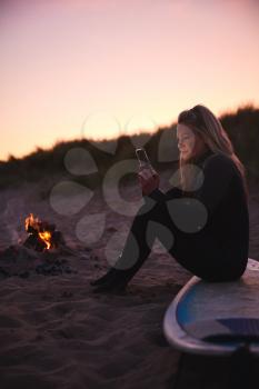 Woman Sitting On Surfboard By Camp Fire On Beach Using Mobile Phone As Sun Sets Behind Her