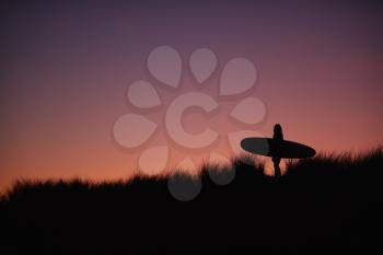 Silhouette Of Female Surfer Carrying Surfboard Across Dunes Against Setting Sun