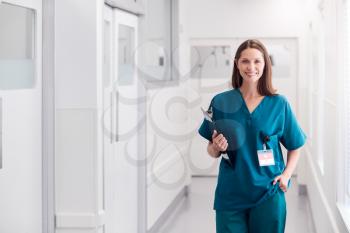 Portrait Of Smiling Female Doctor Wearing Scrubs Standing In Hospital Corridor Holding Clipboard
