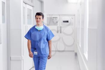 Young Male Doctor Wearing Scrubs Walking Along Hospital Corridor