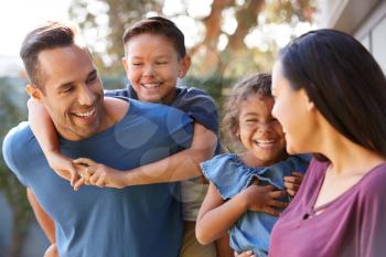 Smiling Hispanic Family With Parents Giving Children Piggyback Rides In Garden At Home
