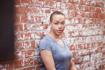 Outdoor Portrait Of Young Woman With Shaved Head Standing In The Street Leaning Against Wall