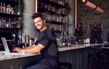 Portrait Of Male Owner Of Restaurant Bar Sitting At Counter Working On Laptop