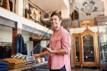 Portrait Of Male Owner Of Fashion Store Using Digital Tablet To Check Stock In Clothing Store