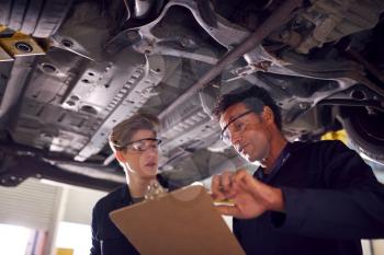 Male Tutor With Student Looking Underneath Car On Hydraulic Ramp On Auto Mechanic Course