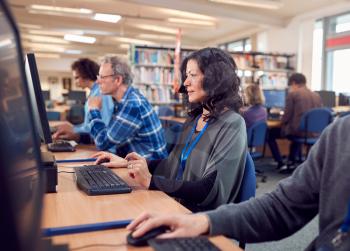 Group Of Mature Adult Students In Class Working At Computers In College Library