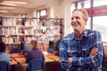 Portrait Of Mature Male Teacher Or Student In Library With Other Students Studying In Background