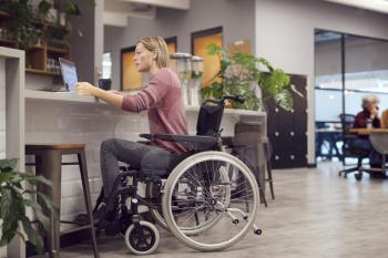 Businesswoman In Wheelchair Working On Laptop In Kitchen Area Of Busy Modern Office