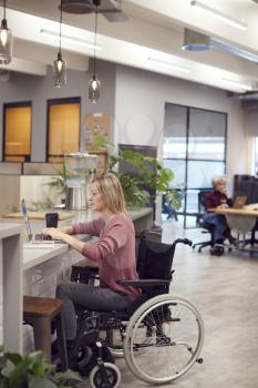 Businesswoman In Wheelchair Working On Laptop In Kitchen Area Of Busy Modern Office