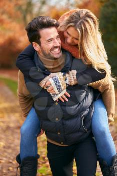 Man Giving Woman Piggyback  As Loving Couple Walking Along Autumn Woodland Path Through Trees
