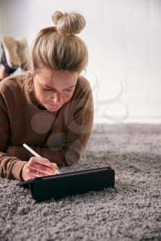 Woman Drawing On Digital Tablet Using Stylus Pen Lying On Carpet At Home