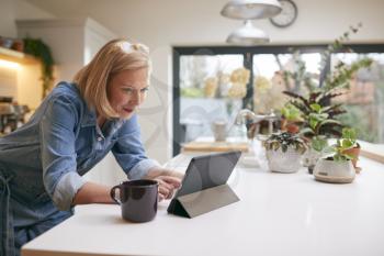 Mature Woman At Home In Kitchen Drinking Coffee And Looking At Digital Tablet