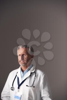 Studio Portrait Of Mature Male Doctor Wearing White Coat Standing Against Grey Background