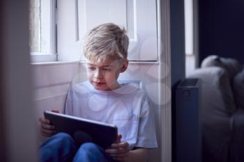 Young Boy Sitting On Window Seat At Home Playing On Digital Tablet