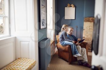 Granddaughter With Grandfather In Chair Looking At Digital Tablet At Home Together