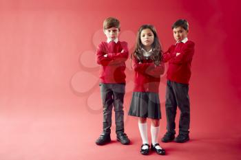 Group Of Elementary School Pupils Wearing Uniform Folding Arms Against Red Studio Background
