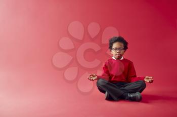Male Elementary School Pupil Wearing Uniform Sitting And Meditating Against Red Studio Background