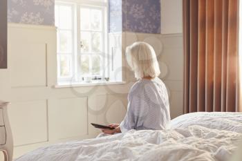 Bereaved Senior Woman Sitting On Edge Of Bed Looking At Photo In Frame