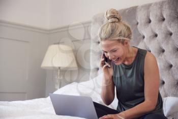 Businesswoman Sitting On Bed With Laptop And Mobile Phone Working From Home During Pandemic Lockdown