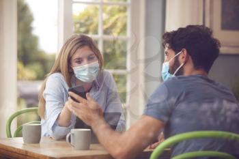 Couple Wearing Masks Meeting In Coffee Shop Looking At Mobile Phone During Health Pandemic Through Window